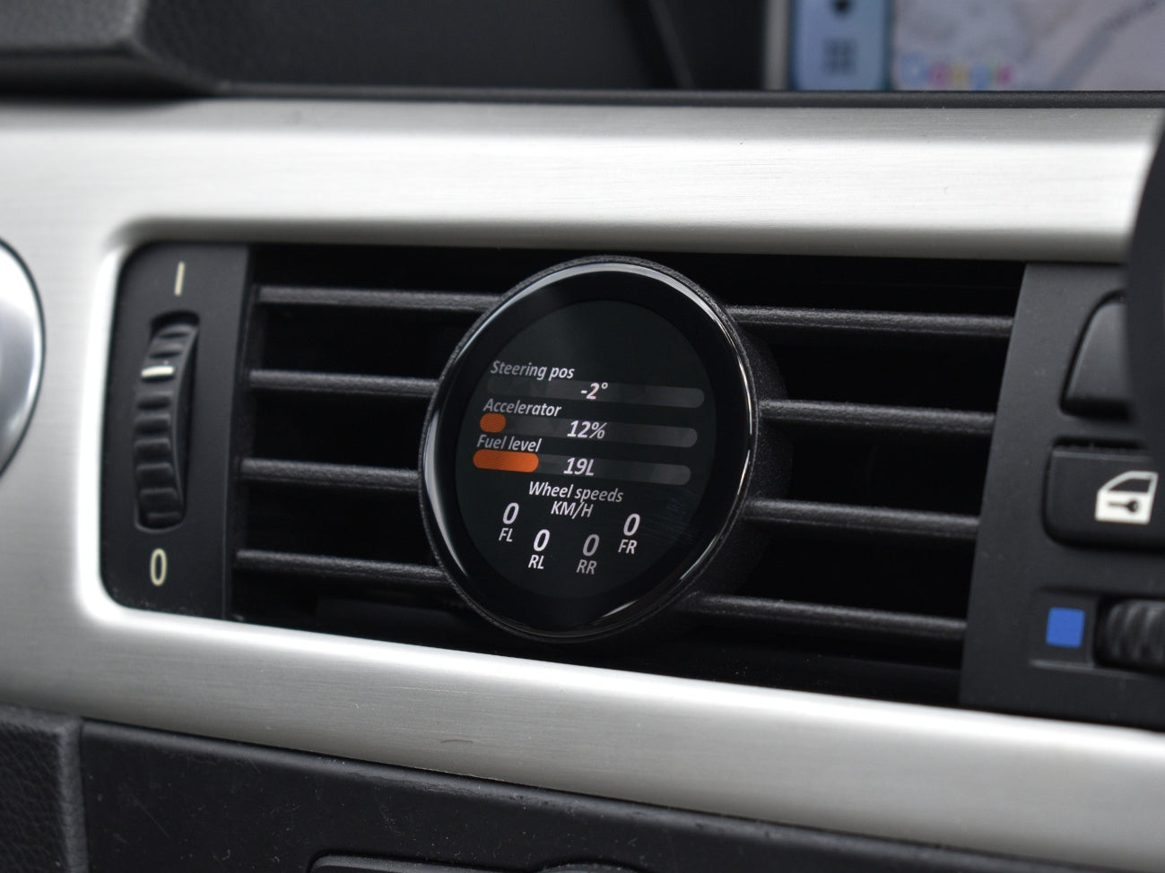 Ein optimaler Alt-Text für dieses Bild wäre:
"Ein COREDISPLAY Datendisplay in einem Lüftungsschlitz, das verschiedene Fahrparameter anzeigt, darunter Lenkwinkel, Gaspedalstellung in Prozent, Tankfüllstand und die Raddrehzahlen der einzelnen Räder.