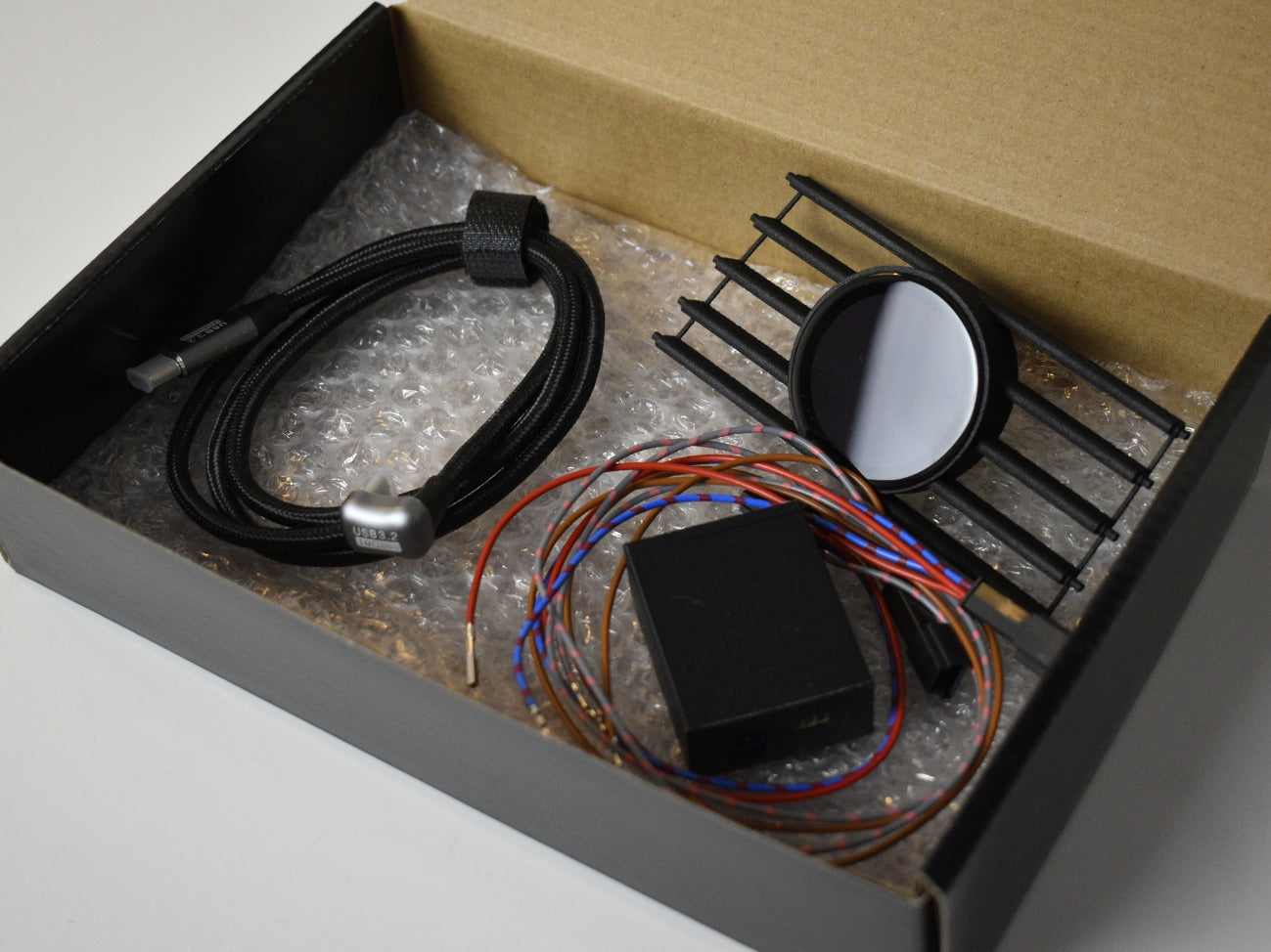 Ein geöffneter Karton mit dem Lieferumfang für das CoreDisplay, bestehend aus dem Lüftungsgitter mit integriertem Display, einem Kabelbaum, einem Steuergerät und einem USB-C-Kabel