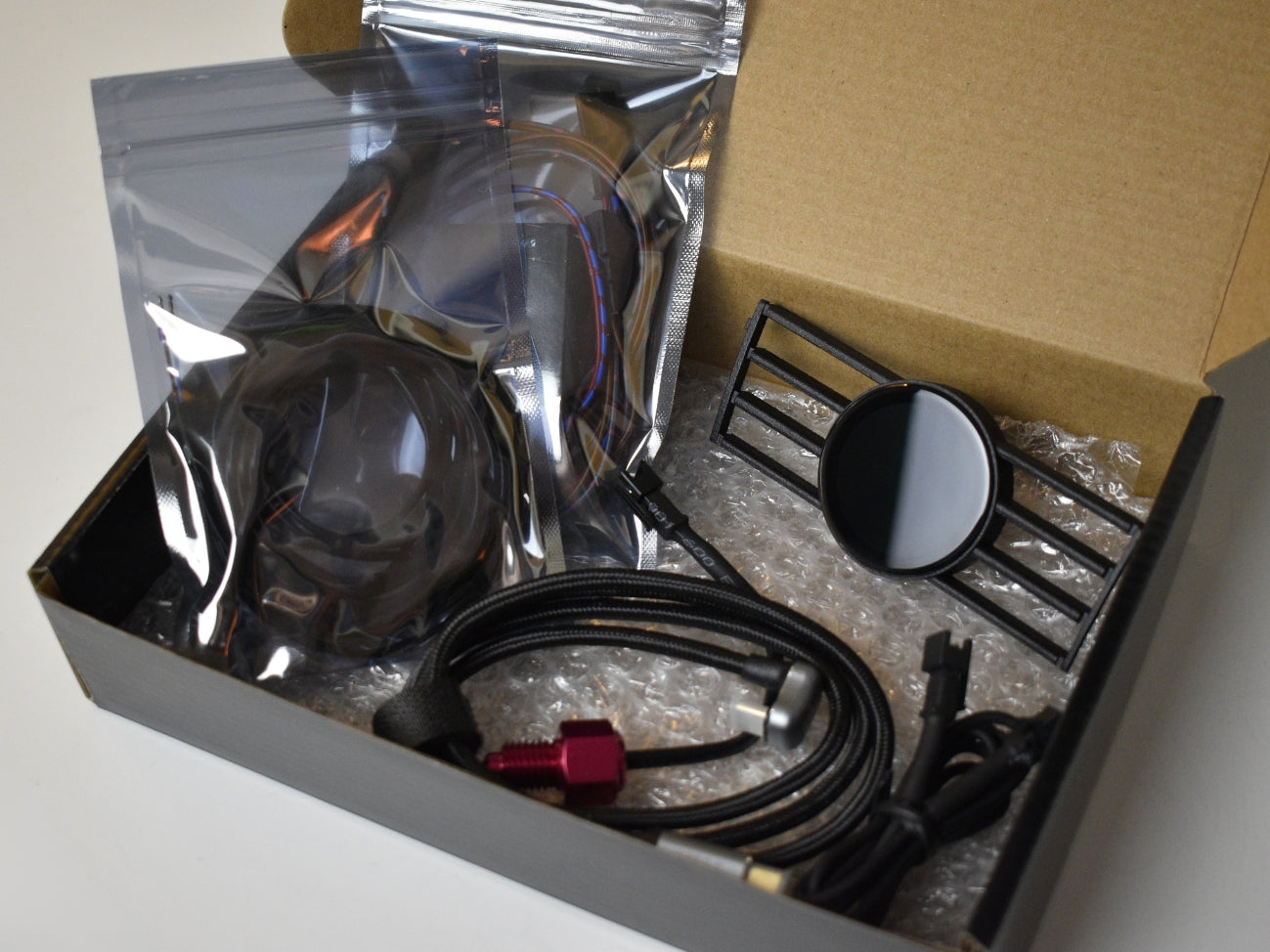 Ein geöffneter Karton mit dem Lieferumfang für das CoreDisplay, bestehend aus dem Lüftungsgitter mit integriertem Display, einem Kabelbaum, einem Steuergerät und einem USB-C-Kabel inkl. Öltemperatursensor