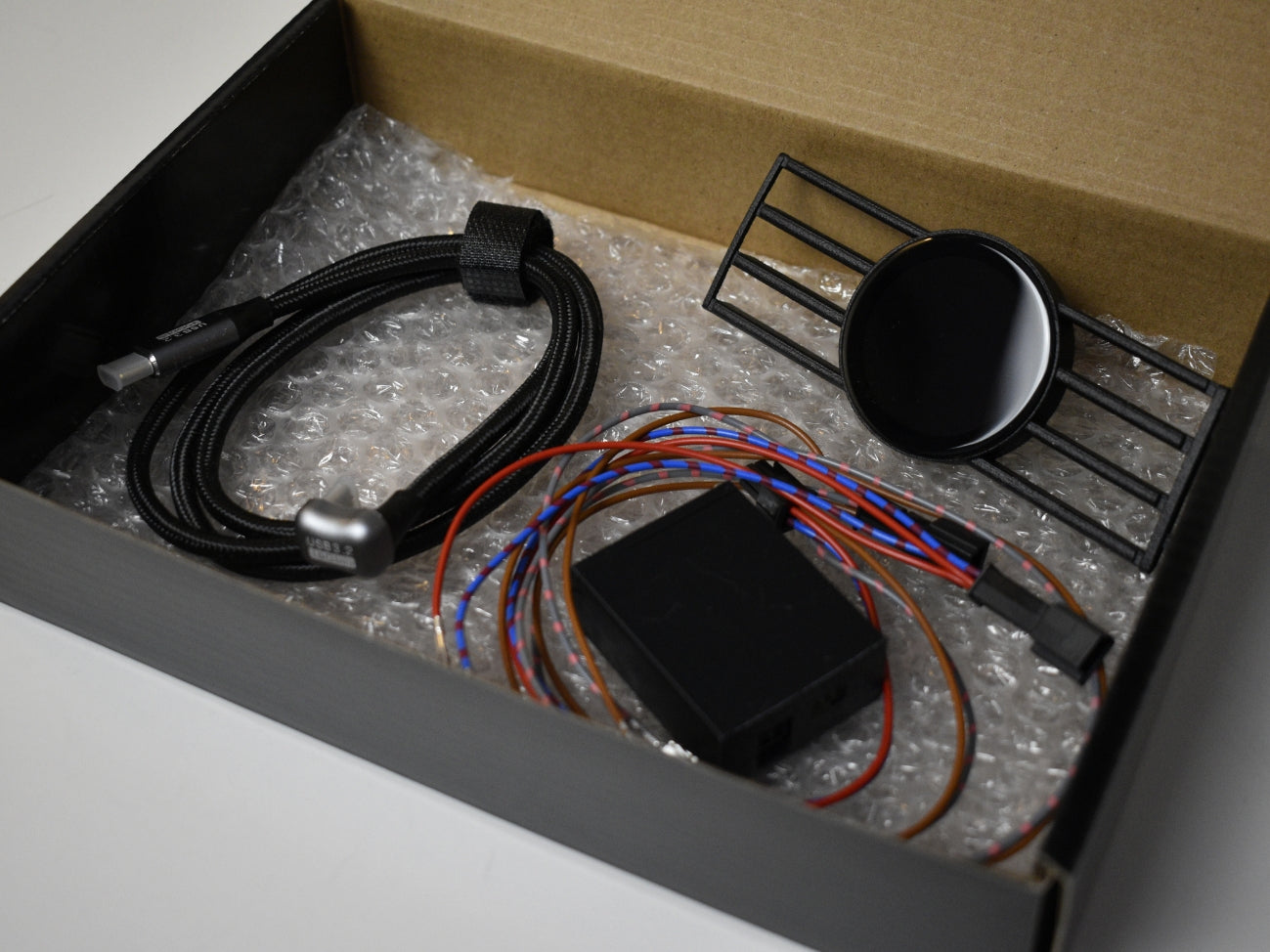 Ein geöffneter Karton mit dem Lieferumfang für das CoreDisplay, bestehend aus dem Lüftungsgitter mit integriertem Display, einem Kabelbaum, einem Steuergerät und einem USB-C-Kabel