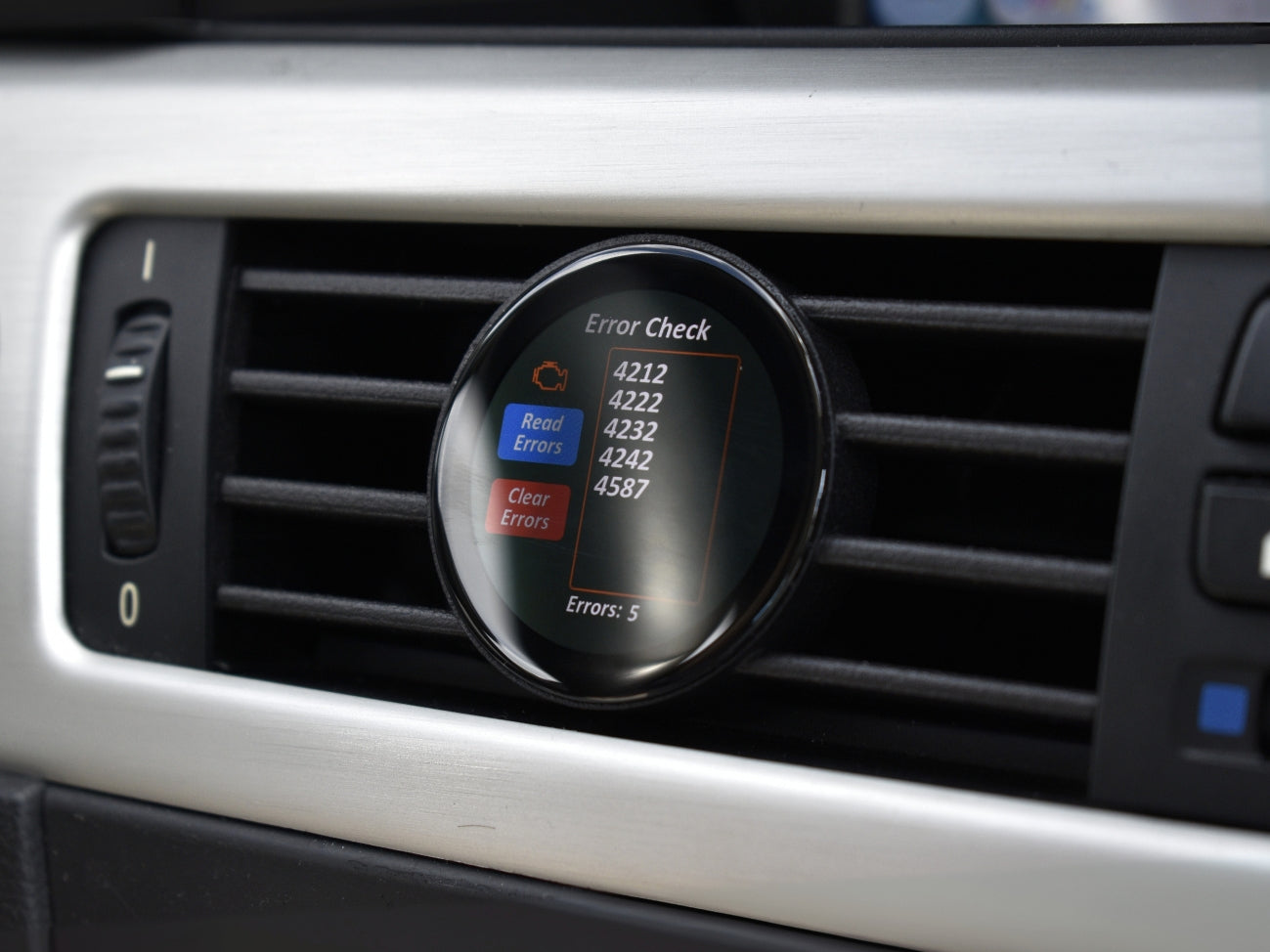 Ein guter Alt-Text für dieses Bild wäre:
"Ein COREDISPLAY Datendisplay, das im Lüftungsschlitz eines Autos montiert ist und den 'Error Check' Modus anzeigt. Das Display zeigt eine Liste von fünf Motorsteuergeräte-Fehlern an und bietet die Option, diese Fehler auszulesen und zu löschen.