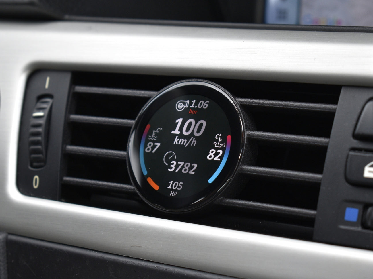 Ein COREDISPLAY GEN3 Datendisplay, eingebaut in den Lüftungsschlitz eines BMW 3er (E9x). Das Display zeigt aktuelle Leistungsdaten wie Geschwindigkeit (100 km/h), Ladedruck (1.06 bar), Kühlmitteltemperatur und die aktuelle PS-Zahl an.