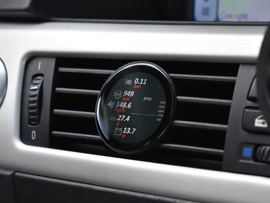 Ein COREDISPLAY Datendisplay, eingebaut in einen Lüftungsschlitz im Auto. Das Display zeigt verschiedene Leistungsdaten an, darunter Ladedruck, Luftmassenstrom in kg/h, G-Kräfte, Drehzahl (RPM) und Batteriespannung.