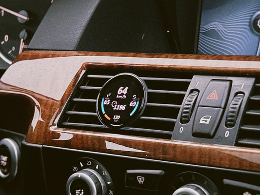 Ein COREDISPLAY Datendisplay, eingebaut in den Lüftungsschlitz eines BMW 5er (E60/E61) mit Holzdekor-Armaturenbrett. Das Display zeigt in Echtzeit Leistungsdaten wie die Geschwindigkeit von 64 km/h an.